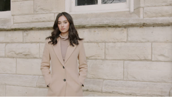 A woman in a camel coat stands against a stone wall, showcasing sleek, polished long hair styled with a gold hair wax stick for a refined fall look.